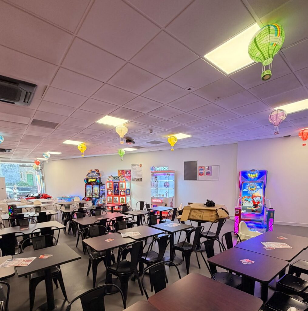 Cafe-style game room with many dark tables and black chairs; arcade machines line the back wall and colorful hot-air balloons hang from the ceiling.